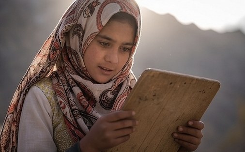 A girl in a patterned headscarf reading from a wooden writing board in golden light